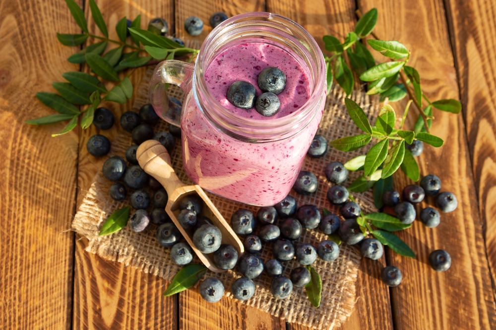 blueberry smoothie in a glass with blueberries on top and blueberry branches on the wooden table with a wooden scoop