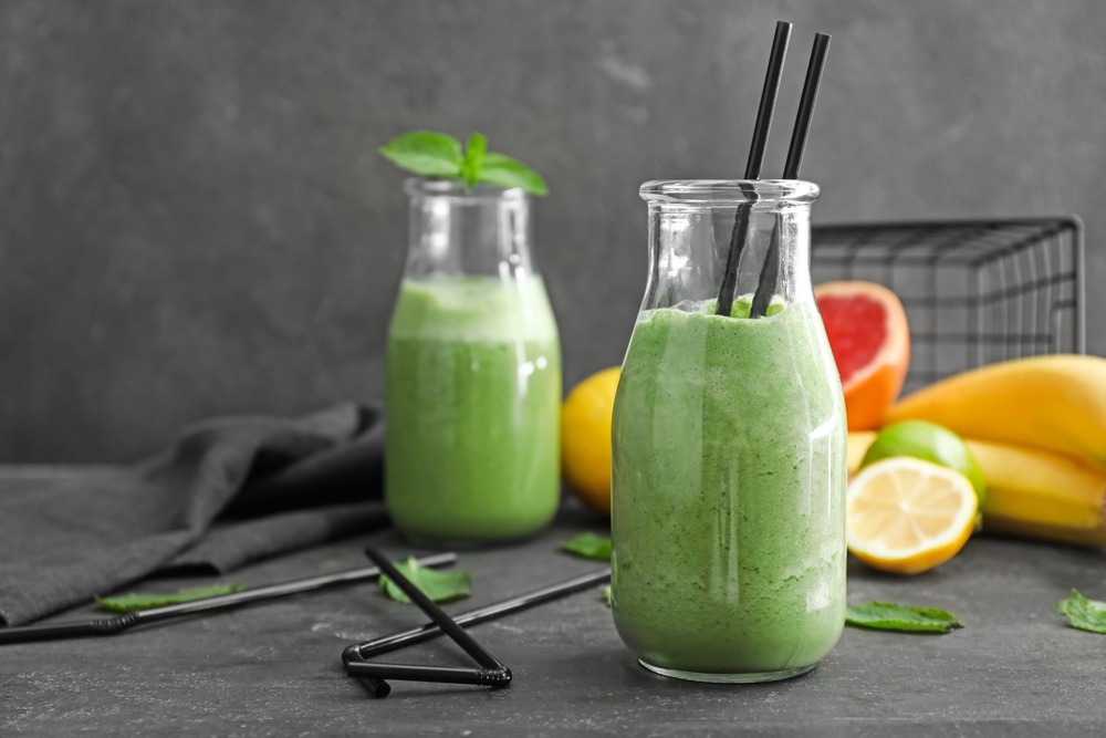 Two green healthy smoothies on a table with basil and straws in and around them with assorted fruit coming out of a basket in the background