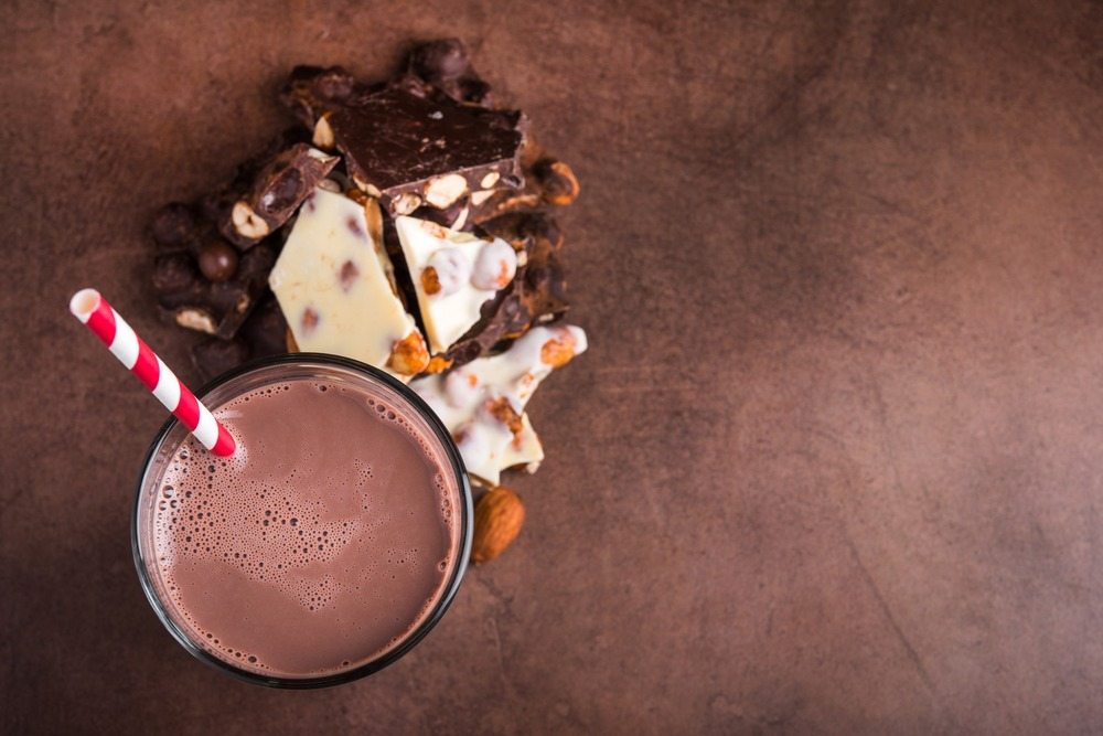 enriched chocolate milk in a glass with a straw with chocolate, nuts, and brittle next to it on the stone countertop