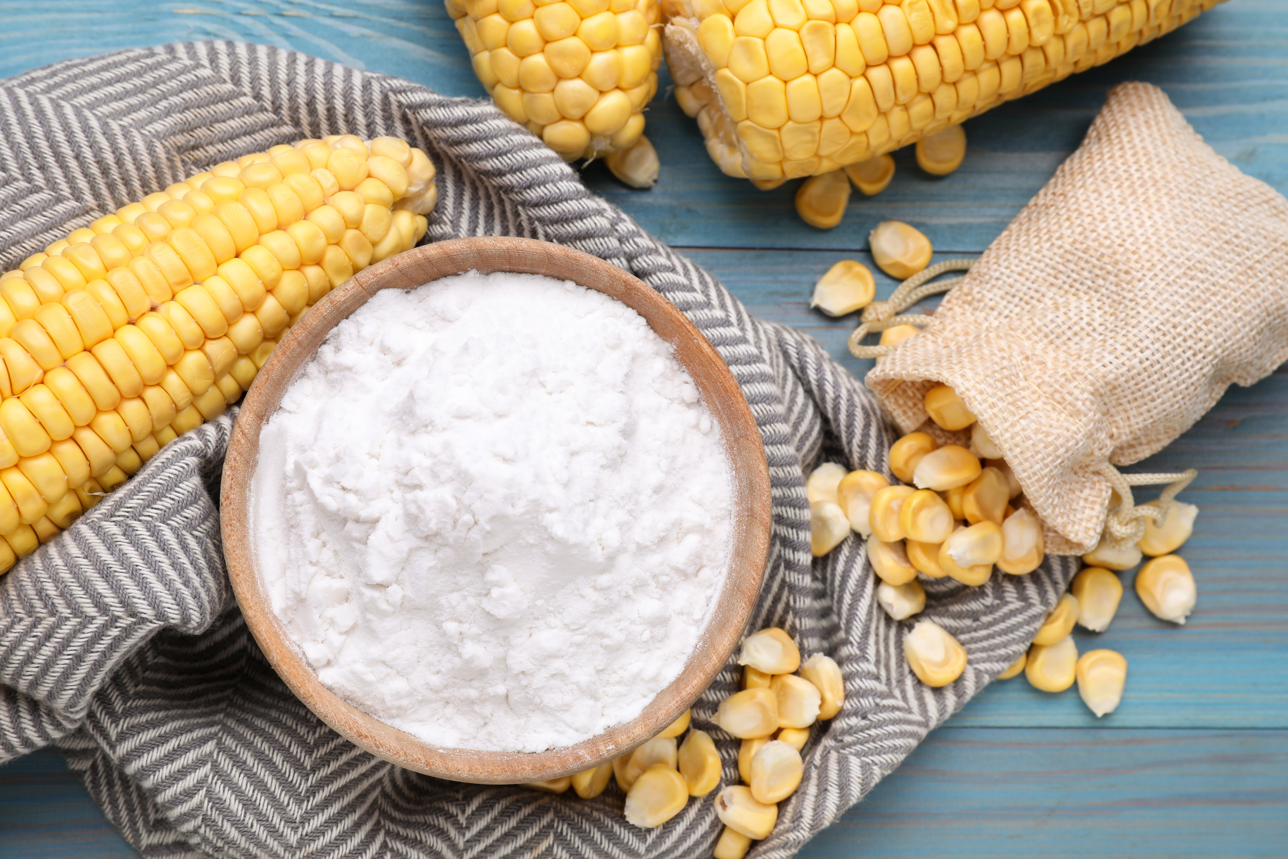 Corn starch in a wooden bowl with corn cobs and a towel on a blue background