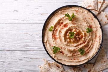 A bowl of fresh hummus sits on a white wooden table