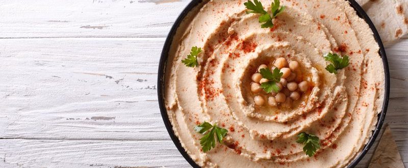 A bowl of fresh hummus sits on a white wooden table