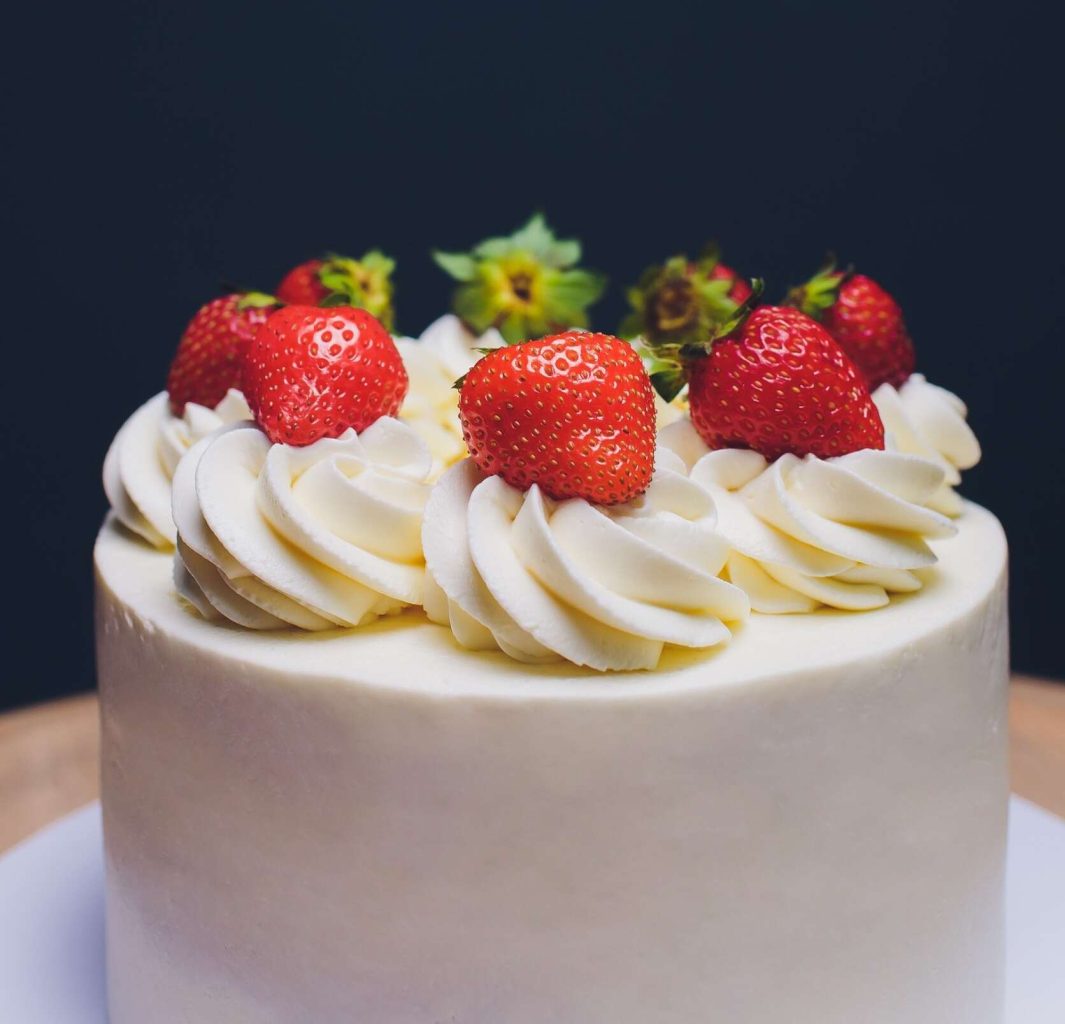 An iced cake topped with strawberries sits on a table with a black background