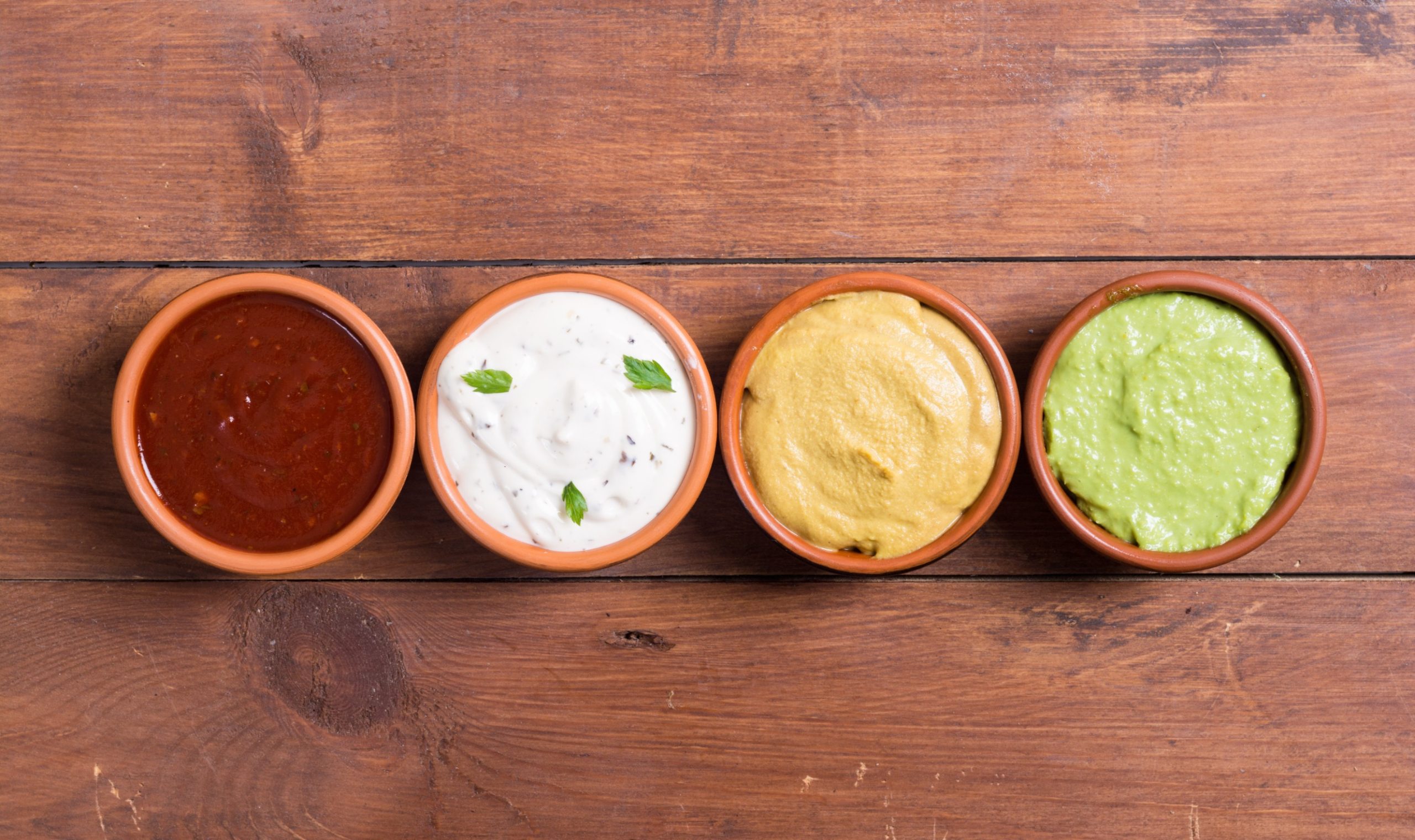 4 different sauces and dips from a sauce ingredient supplier in small bowls atop a wooden table.