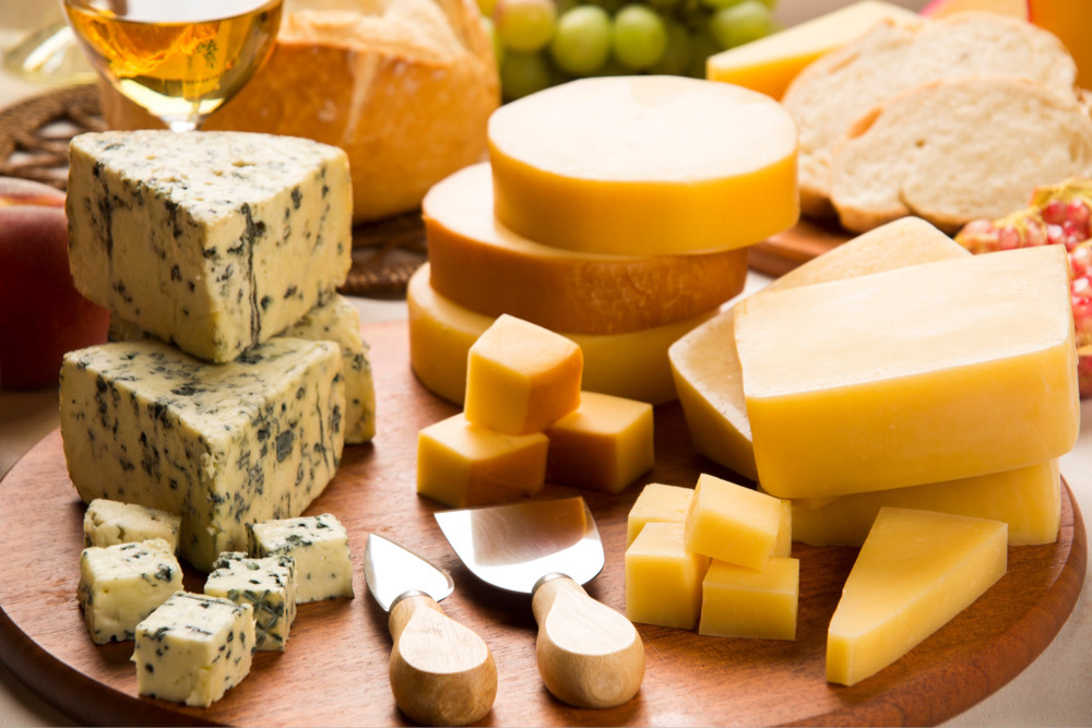 Different kinds of cheese on a large cutting board with two cheese knives nearby from a dairy ingredient supplier